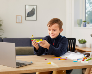 Chłopiec przy stole składa model z lego education i pokazuje do kamerki w komputerze. Ma ekscytację na twarzy.