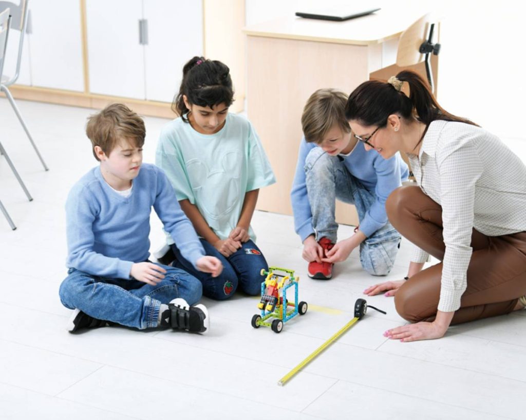 nauczyciel na lekcji fizyki testuje z uczniami model na podłodze i mierzy odległość trasy modelu z klocków lego education
