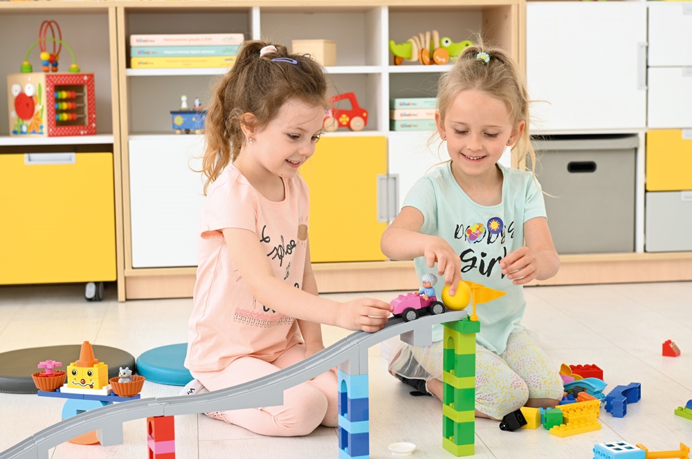 dzieci podczas zajęć w przedszkolu, budują trasę dla pojazdów z klocków lego duplo, dwie dziewczynki, w tle meble przedszkolne żółte